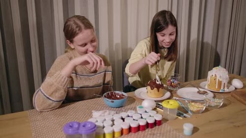 Two Girls Decorating Easter Cakes at a Table Filled with Colorful Sprinkles Paint Jars Brushes and