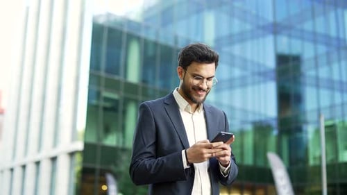 Confident businessman in suit walking near modern glass business office building holding smartphone