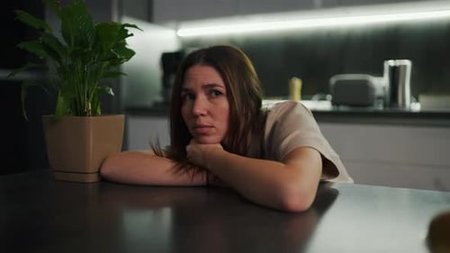 A Sad and Tired Brunette Girl in a Beige Tshirt Sits Leaning on a Black Dining Table Near a Green