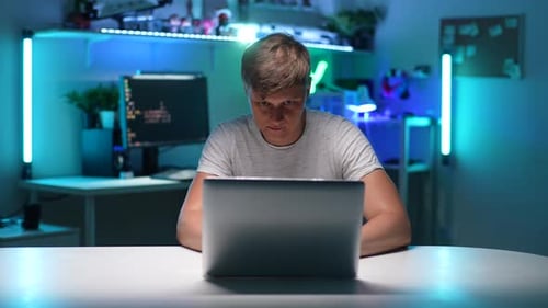 Porträt eines fokussierten jungen Mannes, der im Dunkeln am Tisch sitzt und zu Hause am Laptop arbeitet