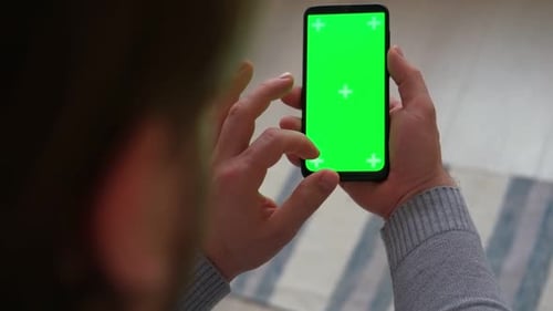 Young Man Sitting at Home Holding Smartphone Green Mockup Screen in Hand Male Person Using Chroma