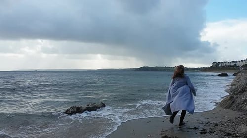 Femme courant sur un rivage venteux avec son long manteau qui virevolte magnifiquement comme des vagues