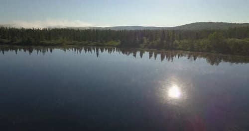 Aerial shot panning left over a calm watered lake with sun reflected facing a beautiful forest in no