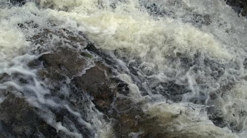 Close-up of turbulent waterfall foam swirling over dark rocks with rapid motion in Ultra slow