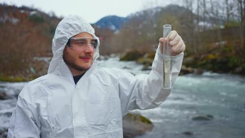 Positive and Smiling Ologist in a Protective White Jumpsuit Tests the Water Quality in the River