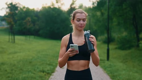 Attractive Sporty Caucasian Runner Woman is Going in the Park Using Smartphone Holding the Sport