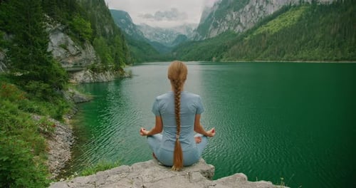 Woman sitting in lotus position by serene lake surrounded by rocky cliffs and trees