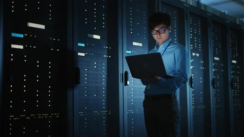 In Dark Data Center: Male IT Specialist Walks along the Row of Operational Server Racks, Uses Lapto