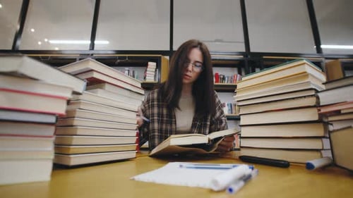 Young Woman Studying in a Library Surrounded By Stacks of Books While Taking Notes at a Wooden Table