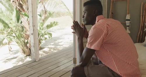 Thoughtful african american man sitting in doorway of beach house looking away, slow motion