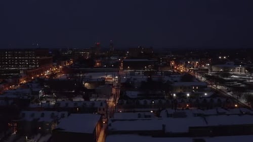 Rising aerial of urban city in USA during dark winter night. Snow covers rooftops. Snowy cold establ