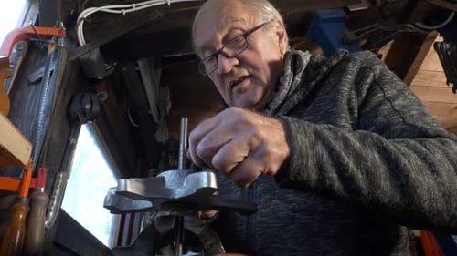 Senior caucasian worker uses hand file to work on a metal piece in a small dark workshop