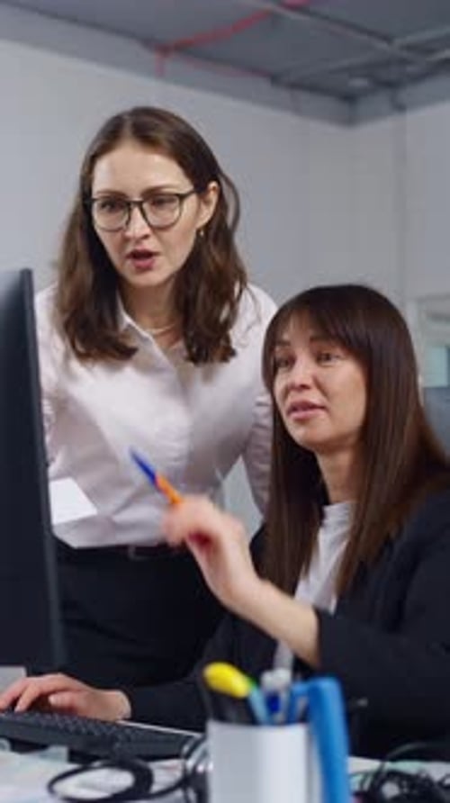 Two Professional Women Collaborating at Computer Workstation in Modern Office Discussing Project