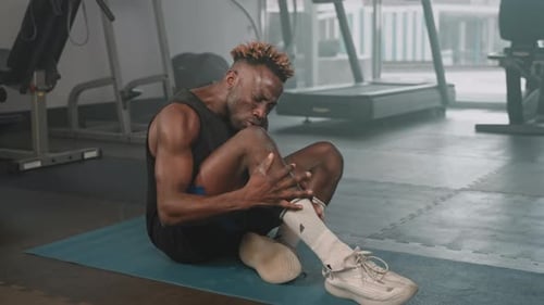 Foot Cramp After Sport Workout Black Athlete is Rubbing His Leg Sitting on Floor in Gym