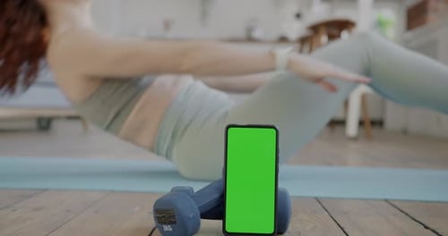 Closeup of Smartphone with Chroma Key Green Screen and Young Woman Doing Sports on Yoga Mat at Home