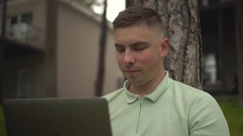 A Young Man Sits Under a Tree and Works on a Laptop A Man with a Laptop in Nature Sitting on the
