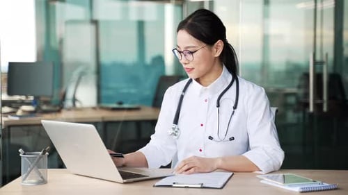 Portrait of young smiling asian female doctor in white coat working on laptop in hospital clinic