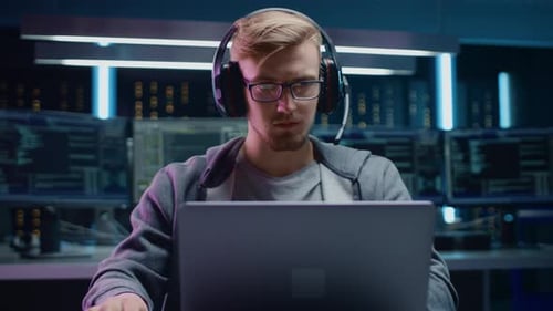 Portrait of Software Developer / Hacker / Gamer Wearing Glasses and Headset Sitting at His Desk and
