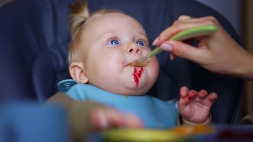 Lovely blue-eyed baby with a bib is fed from spoon. Close up. Nourishing an infant at home.