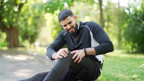 Bearded handsome male runner experiences muscle pain and discomfort while sitting in city park.