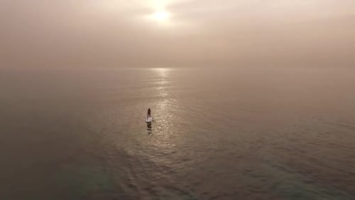 Aerial pull back of one stand up paddleboarder in ocean to reveal kneeling paddler at sunset