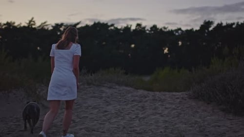 Attractive young woman and her dog walking up hill in sand dunes at golden hour