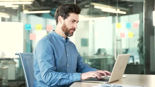 Side view. Focused businessman typing on a laptop sitting at a workplace in office. Bearded manager