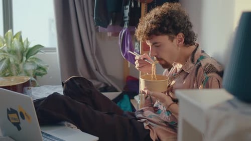 Young Adult Eats Noodles on Bed with Laptop
