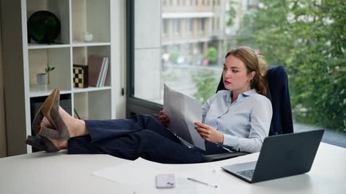 in a Modern Office with a Large Window a Beautiful Worker in a Blue Shirt Sits with Her Feet on the