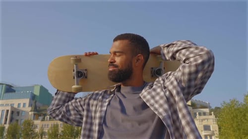 Portrait of Cool Black Man with Skateboard on Shoulders Relaxing in City Street