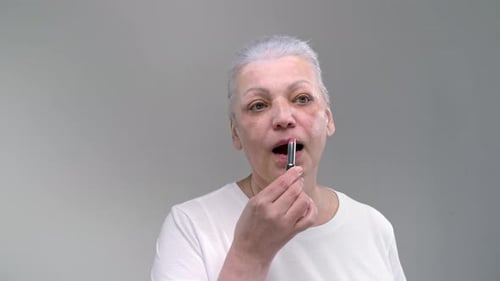 Concentrated Mature Woman with Gray Hair Applies Pink Color Lipstick Standing on Light Background