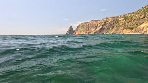 Sea Water Surface Camera Flies Over the Calm Azure Sea with Volcanic Rocky Shores on Background
