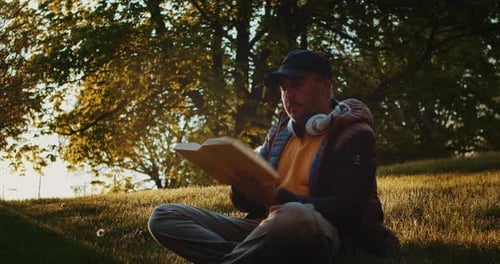 Mann liest ein Buch auf einer Lichtung im Park mit Kopfhörern