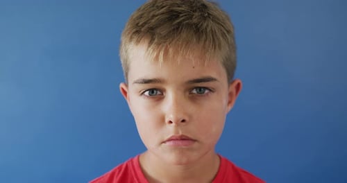 Close up of portrait of sad caucasian boy looking at camera on blue background, slow motion
