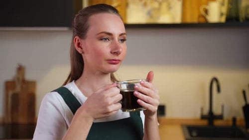 Pretty Young Woman in a Green Kitchen Apron with a Cup of Coffee in Her Hands is Sitting in the