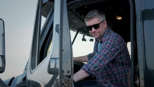 Close Up of Portrait of Professional Truck Driver Sits in His Truck and Behind Him Parked Long Haul