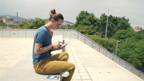 Woman Browsing Internet On Tablet Sitting On Bench In City