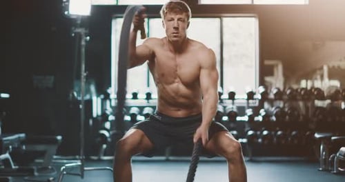 Attractive muscular man working out in the gym using battle ropes with high intensity,