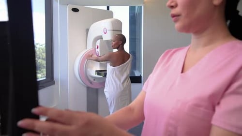 Female patient in gown holding mammogram at clinic while female tech tapping console starting scan