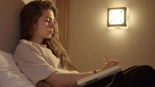 Attractive Young Woman in White Tshirt Writes Entries in Diary or Notebook While Lying on Bed with