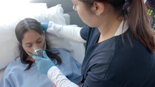 Diverse female doctor putting oxygen mask on female patient in hospital bed, slow motion