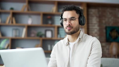 Focused IT specialist in wireless headphones works remotely on a laptop sitting on a sofa in room