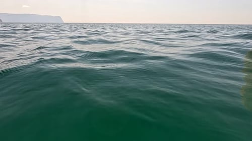 Sea Water Surface Camera Flies Over the Calm Azure Sea with Volcanic Rocky Shores on Background