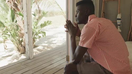 Thoughtful african american man sitting in doorway of beach house looking away, slow motion