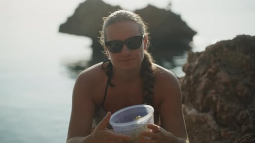 A young woman in a swimsuit is sitting on rocks in the sea. There is a rock in the water behind. She