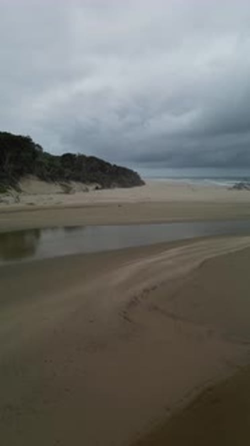 Aerial reverse over a serene beach with a winding river meeting the ocean on an overcast day.