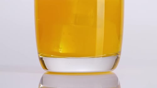 Close Up Of Orange Juice Being Poured Into A Glass With Ice Cubes On The Light White Background