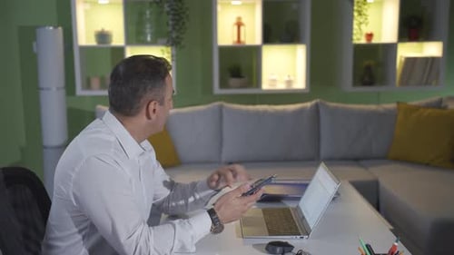 Man Uses Laptop, Smiling at Smartphone at Desk