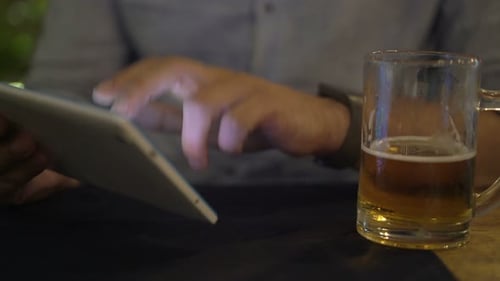Young man browsing the internet on tablet computer and drinking beer at night in cafe