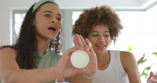 Duas mulheres africanas mostrando creme na cozinha, jarra estendida para a esquerda, bombeando para a direita, ambas testando textura